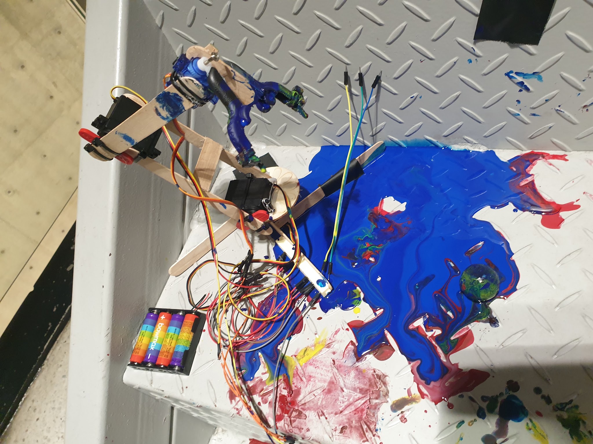 Paint-covered robotic arm beside its wired mechanism, ball, and painted surface